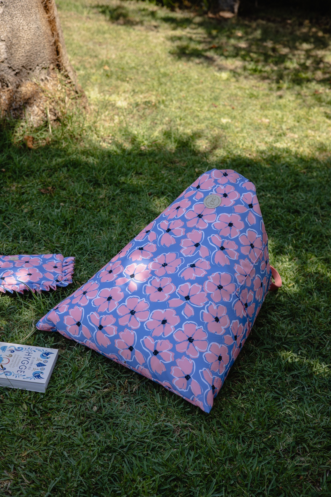 Pufe triangular exterior com padrão floral rosa e lilás na relva junto a livro e bolsa