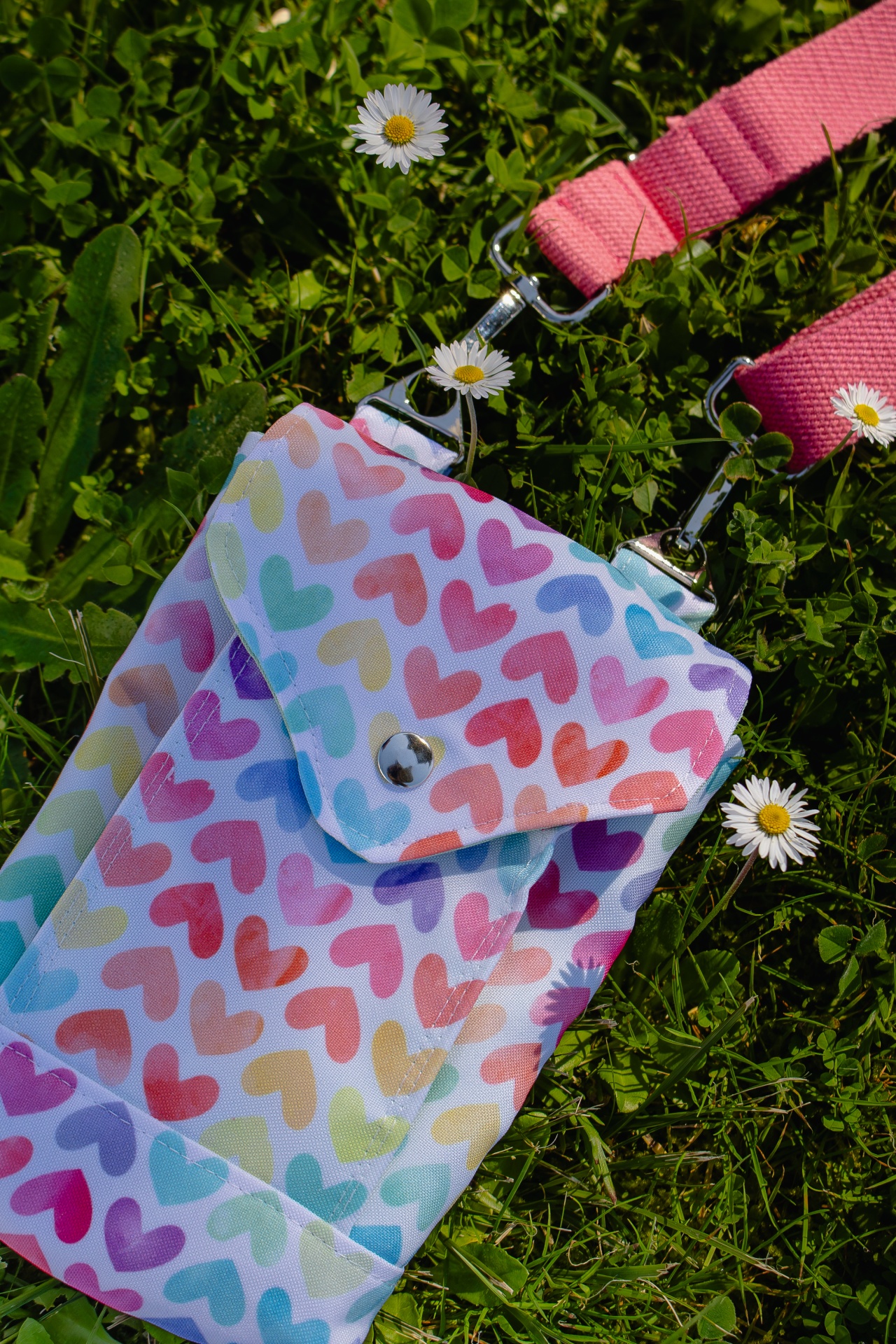 Bolsa colorida com padrão de corações e alça rosa sobre relva verde com margaridas.