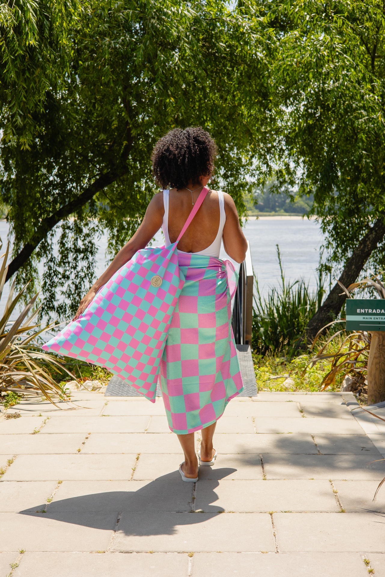 Mulher com fato de banho branco, toalha e saco aos quadrados rosa e azul junto a lago com vegetação.
