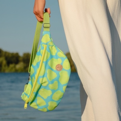Mala azul e verde com padrão segurada por uma mão, próxima a calças brancas, céu azul e vegetação ao fundo