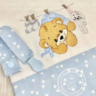 Fralda branca e azul claro com bordado de ursinho e coelho, padrão de triângulos, presilha para chupeta, etiqueta com texto Handmade by Os Babadides Coloridas da Célia