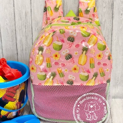 Mochila rosa com padrão tropical e bolso de rede, sobre chão de madeira clara com brinquedos azuis e balde colorido