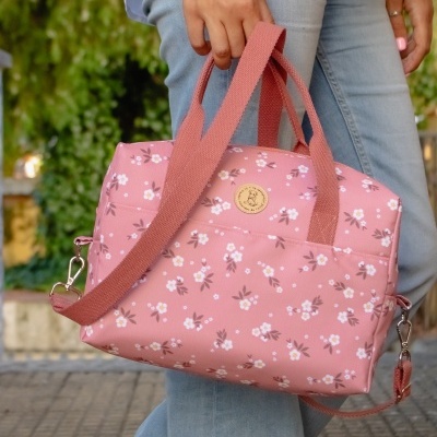 Mala rosa com flores brancas e alças rosa, em mãos de pessoa de jeans azul e camisa branca.