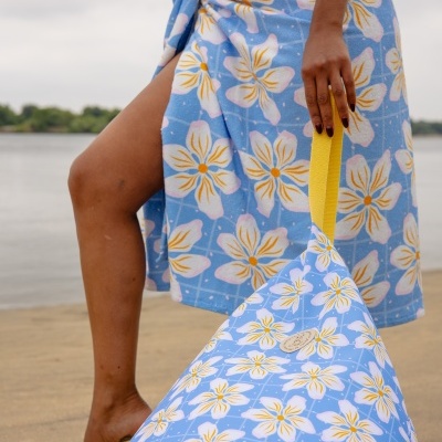 Toalha de praia azul com padrão floral amarrada na cintura e saco com padrão floral e alça amarela na praia