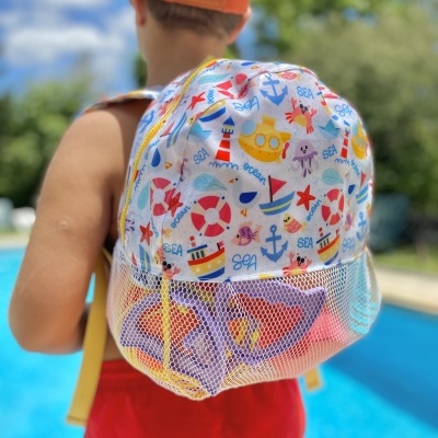 Mochila infantil colorida com padrão marinho e parte inferior em tela, vista nas costas de uma criança junto a uma piscina