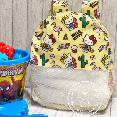 Mochila infantil amarela com padrão da Hello Kitty com cactos, flamingos e cupcake ao lado de brinquedos e copo do Spider-Man