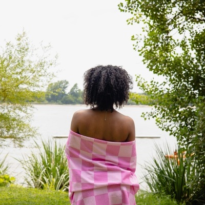 Pessoa de costas com toalha rosa xadrez ao ar livre junto a um lago e árvores