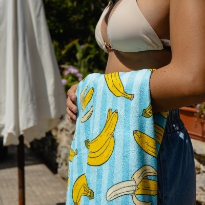 Toalha de banho azul com bananas amarelas segurada por pessoa com fato de banho bege ao ar livre