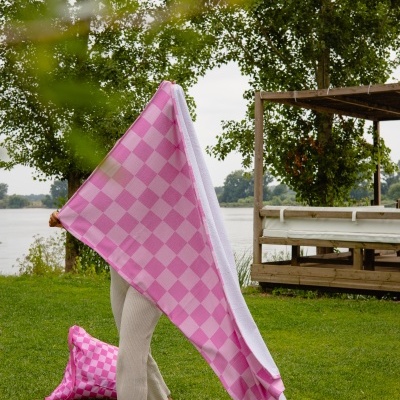 Pessoa segurando uma toalha rosa quadriculada ao ar livre numa área verde junto a um lago