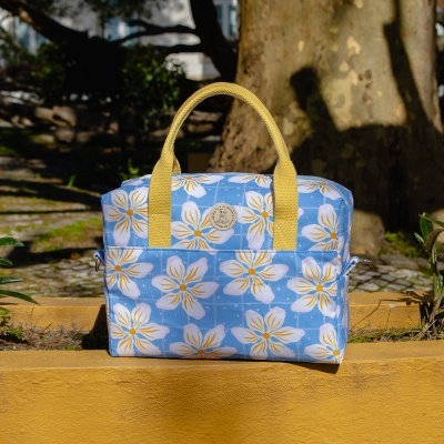 Mala de mão azul com flores brancas e amarelas, alças amarelas, etiqueta redonda, muro amarelo ao fundo