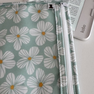 Necessaire azul claro com flores brancas e fecho branco sobre leitor Kobo com texto em português