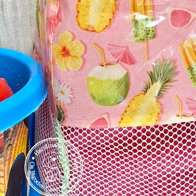 Mochila rosa com padrão tropical e rede branca ao lado de balde azul com brinquedos