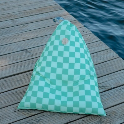 Pufe triangular verde com padrão de quadrados branco e etiqueta circular de couro, posto em deque de madeira junto à água