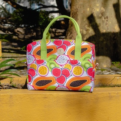 Mala de mão com padrão de frutas tropicais e alças verdes sobre muro amarelo