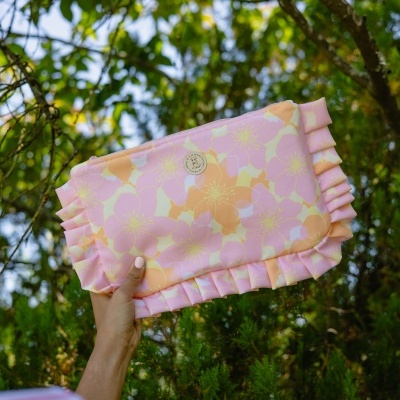 Bolsa tipo clutch rosa com padrão floral e detalhe franzido, segurada à mão em ambiente natural de árvores