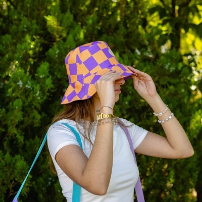 Pessoa com chapéu bucket laranja e roxo em padrão xadrez, t-shirt branca, pulseiras, com fundo verde de árvores