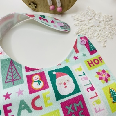 Babete colorido de Natal com imagens e palavras festivas, fecho de pressão cor-de-rosa, sobre fundo branco com decoração natalícia