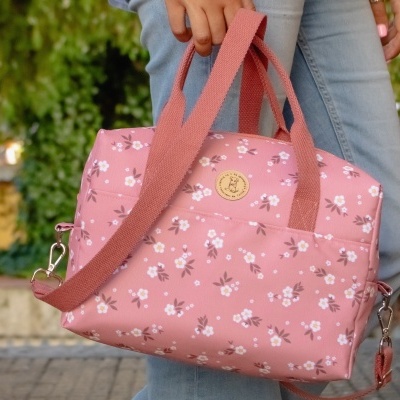 Mala rosa com padrão floral segurada por uma mão em fundo exterior.