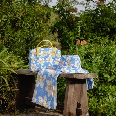 Mala, chapéu e toalha com padrão floral azul claro, branco e amarelo sobre banco de madeira rústico.