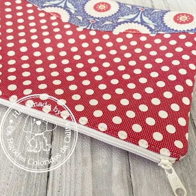 Estojo vermelho com bolinhas brancas e fecho branco em tecido, com detalhe azul floral e selo branco circular com elefante.