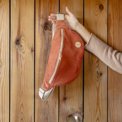 Mochila terracota em tecido canelado com fecho dourado e alças largas pretas e cinzentas segurada por uma mão