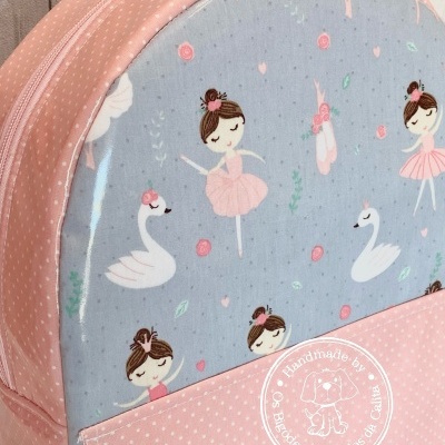 Mochila infantil com padrão de bailarinas e cisnes em fundo azul claro e detalhes rosa com bolinhas