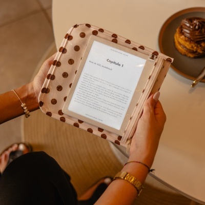 Capa para leitor digital bege com bolinhas castanhas, e-reader dentro a mostrar texto, mãos a segurá-lo junto a uma mesa com bolo e caixa de lenços.