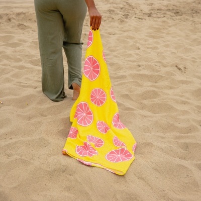 Pessoa a segurar uma toalha amarela com padrão de fatias de citrinos enquanto está na praia.