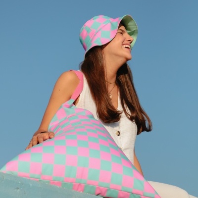 Mulher sorridente a usar chapéu de balde com padrão quadriculado em rosa e verde, segurando almofada com o mesmo padrão, céu azul de fundo.