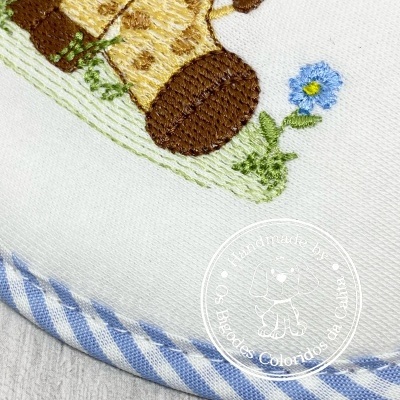 Produto têxtil branco com bordado de animal e flor, fita azul às riscas e selo branco redondo