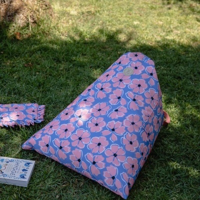 Pufe triangular exterior com padrão floral rosa e lilás na relva junto a livro e bolsa