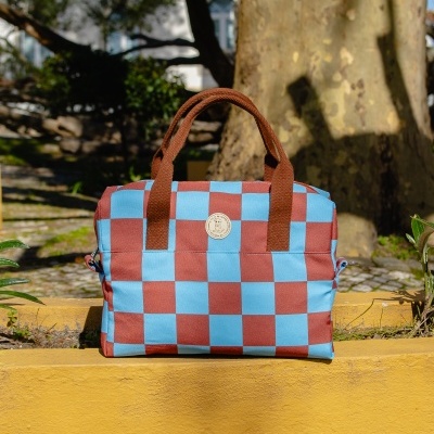 Mala azul e castanha quadriculada com alças castanhas num muro amarelo com árvores atrás