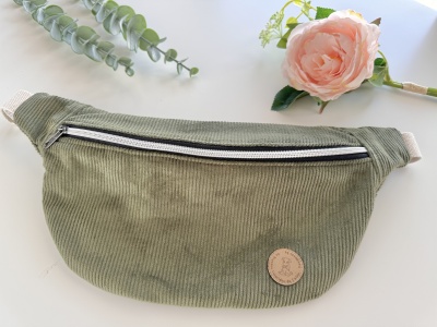 Pochete verde com fecho de correr e etiqueta redonda sobre mesa branca com flor e folhas.