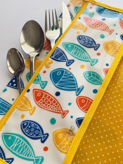 Guardanapo amarelo com pontos brancos e padrão de peixes coloridos junto a talheres metálicos.