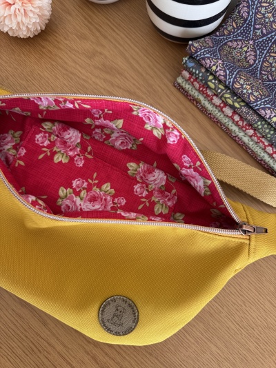 Bolsa de cintura amarela com interior floral vermelho aberta sobre mesa de madeira junto a vaso e tecidos dobrados