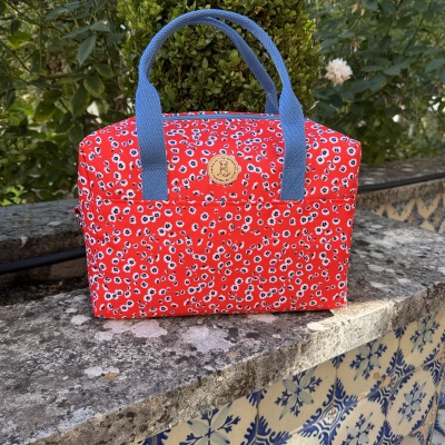 Bolsa vermelha com padrão floral, alças azuis e etiqueta redonda castanha em superfície de pedra com azulejos.