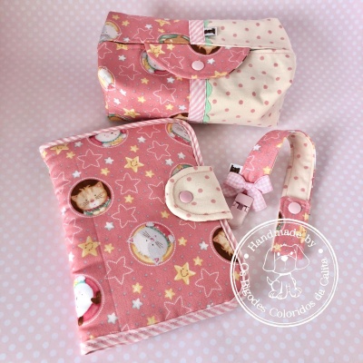 Acessórios em tecido rosa com padrão de gatos e estrelas, incluindo bolsa, carteira e alça, sobre fundo rosa com bolinhas.