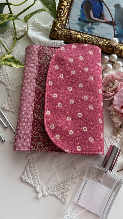 Carteira de tecido rosa com padrão floral sobre toalha branca bordada, frasco de vidro e moldura dourada