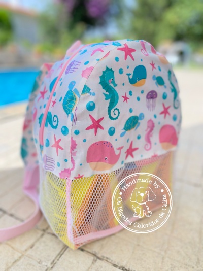 Mochila infantil rosa com padrão marinho colorido e bolso de rede amarelo em área exterior junto a piscina
