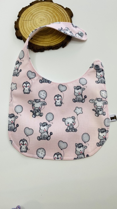 Babete rosa com padrão de animais bebés e balões, com botão de pressão, sobre placa de madeira