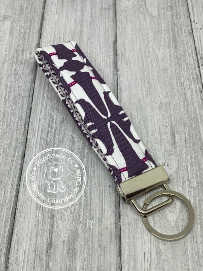 Porta-chaves em tecido branco e roxo com argola metálica sobre fundo de madeira cinzenta