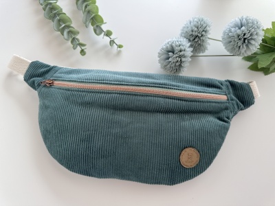 Pochete verde-azulado em veludo cotelê com fecho de correr bege e etiqueta redonda castanha, decorada com folhas e flores cinzentas.