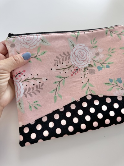 Necessaire rosa com flores e bolinhas pretas, fechada com zip, segurada por mão.