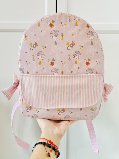 Mochila rosa infantil com padrão de bailarinas e flores, bolso frontal riscado e alças cor-de-rosa.