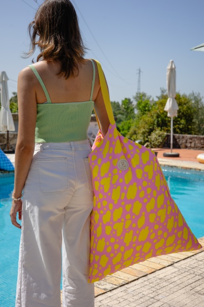 Mulher com top verde claro e calças brancas junto a piscina com bolsa rosa e amarelo.