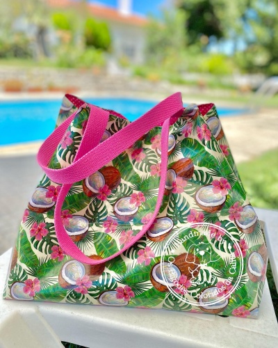 Saco de tecido estampado com cocos, folhas verdes e flores, com alças rosas, ao lado de uma piscina.
