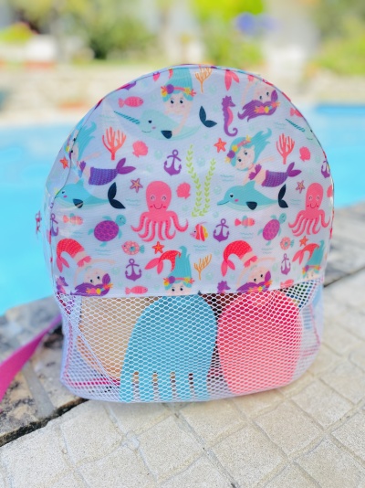Mochila infantil colorida com padrão de sereias e animais marinhos, com bolso frontal de rede contendo brinquedos de praia