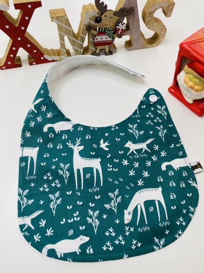 Babete de bebé azul petróleo com padrão branco de animais e plantas, decorado com motivos natalícios ao fundo.