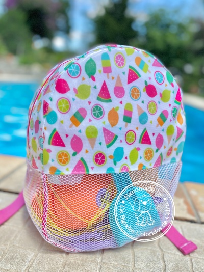 Mochila infantil colorida com padrão de frutos e gelados, parte inferior em rede branca, perto de piscina