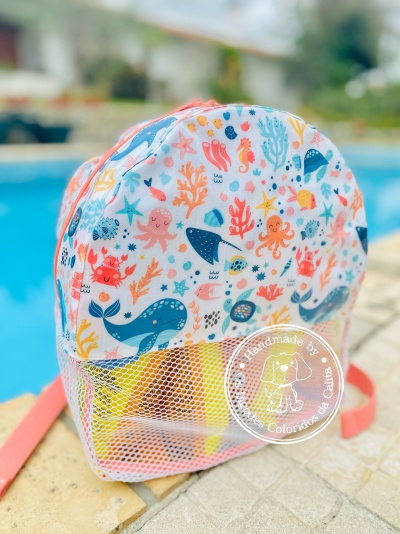 Mochila infantil colorida com padrão marinho e bolso de rede branca, junto a piscina